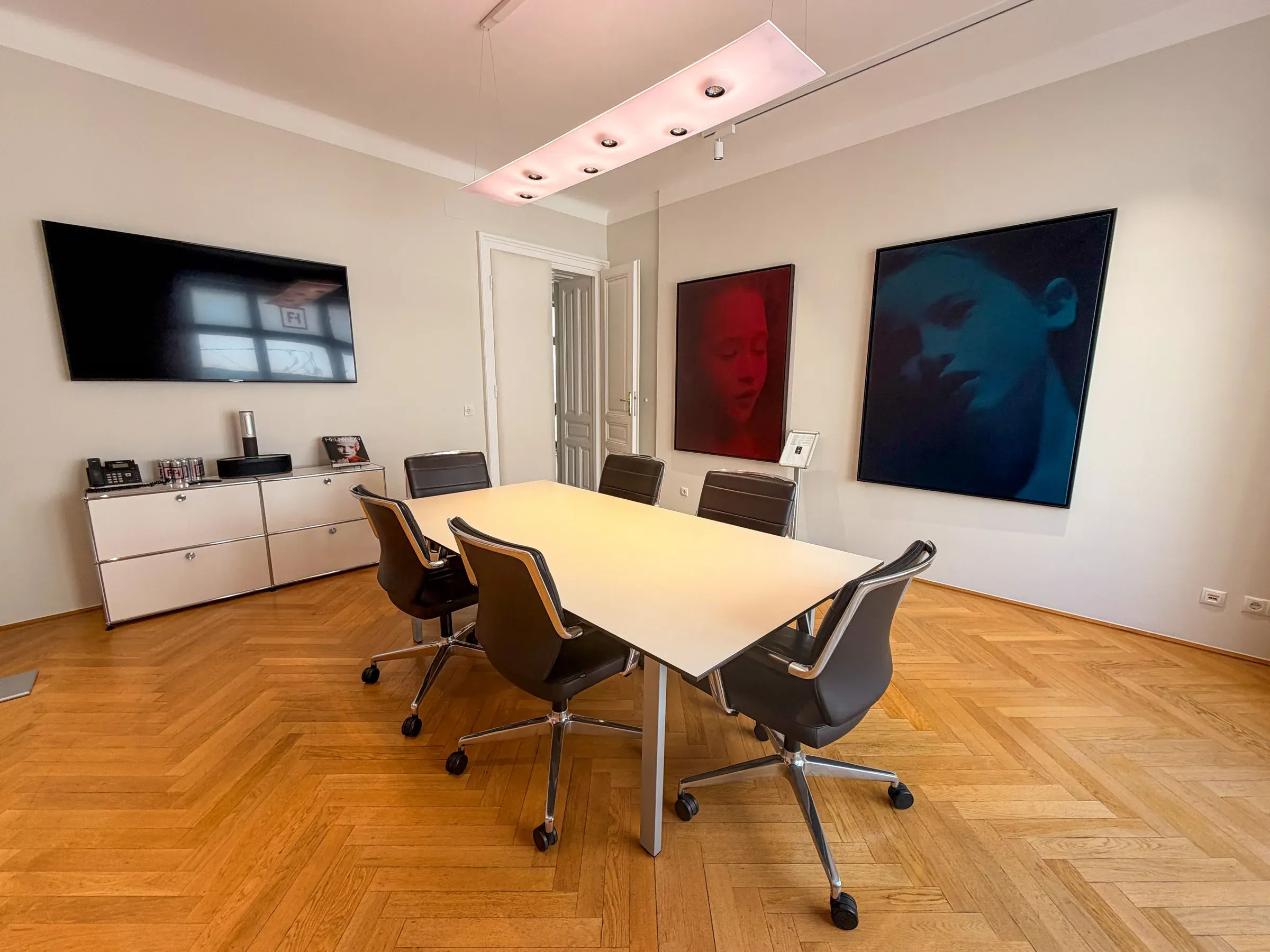 Besprechungsraum mit langem Tisch, Fischgrätparkett und zwei großformatigen Porträts an der Wand.